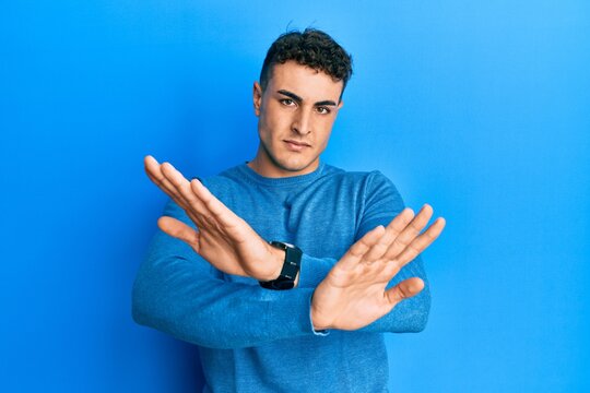 Hispanic young man wearing casual winter sweater rejection expression crossing arms doing negative sign, angry face