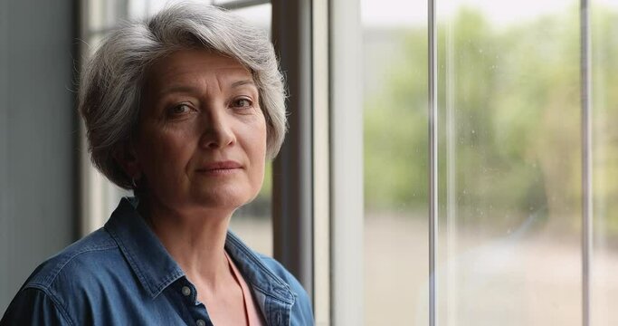Close up view middle-aged 55s grey-haired thoughtful female standing alone indoor looking out the window. Attractive woman with short hairstyle smiling staring at camera having attractive appearance