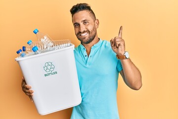 Handsome man with beard holding recycling wastebasket with plastic bottles surprised with an idea...