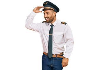 Handsome man with beard wearing airplane pilot uniform very happy and smiling looking far away with hand over head. searching concept.