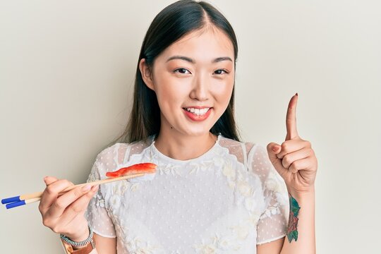 Young chinese woman eating prawn sushi using chopsticks smiling with an idea or question pointing finger with happy face, number one
