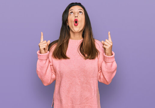 Beautiful hispanic woman wearing casual winter sweater amazed and surprised looking up and pointing with fingers and raised arms.
