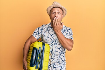 Handsome muscle man holding suitcase going on summer vacation covering mouth with hand, shocked and...
