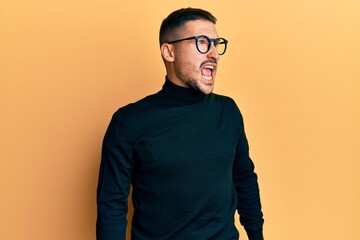 Obraz premium Handsome man with tattoos wearing turtleneck sweater and glasses angry and mad screaming frustrated and furious, shouting with anger. rage and aggressive concept.