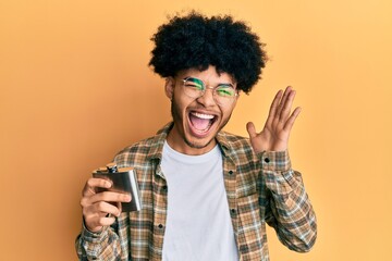 Young african american man with afro hair drinking alcohol from whiskey flask screaming proud, celebrating victory and success very excited with raised arms