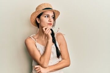 Young hispanic woman wearing summer hat thinking concentrated about doubt with finger on chin and...