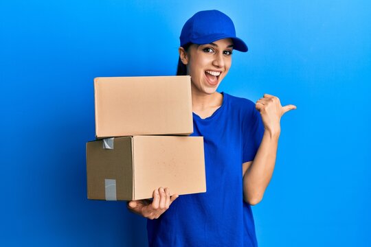 Young hispanic woman holding delivery package pointing thumb up to the side smiling happy with open mouth