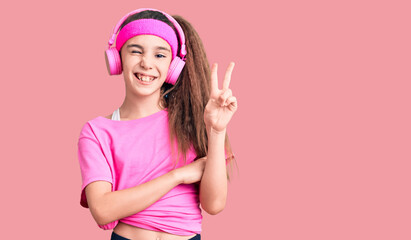Obraz na płótnie Canvas Cute hispanic child girl wearing gym clothes and using headphones smiling with happy face winking at the camera doing victory sign. number two.