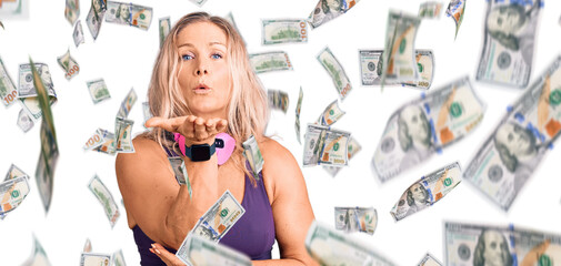 Middle age fit blonde woman wearing gym clothes and using headphones looking at the camera blowing...