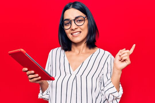 Young beautiful latin woman holding touchpad smiling happy pointing with hand and finger to the side