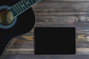 Fototapeta premium A black guitar and a black tablet screen on a brown wooden surface close-up. The concept of a layout for advertising online guitar courses.