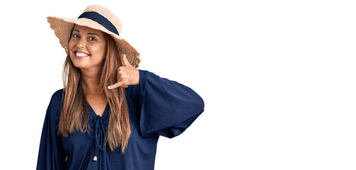 Middle age hispanic woman wearing summer hat smiling doing phone gesture with hand and fingers like...