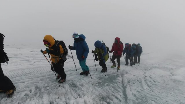 Mountaineering activity on the steep mountain slope. Clip. Group of extreme tourists walking one by one on icy surface of a hill in foggy cold weather.