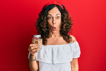 Middle age hispanic woman holding jar with walnuts scared and amazed with open mouth for surprise, disbelief face