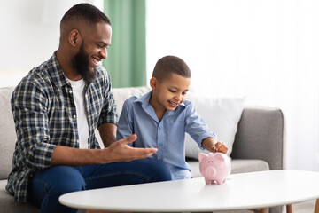Obraz na płótnie Canvas African Father And Son Putting Personal Savings In Piggybank Indoor