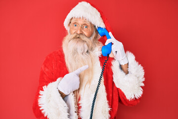 Old senior man wearing santa claus costume using vintage telephone smiling happy pointing with hand and finger