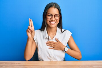 Beautiful hispanic woman wearing casual clothes sitting on the table smiling swearing with hand on...