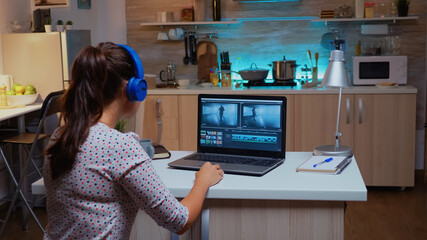 Fototapeta na wymiar Professional colorist working in video footage during post production. Videographer editing audio film montage on modern device, laptop sitting on desk in modern kitchen in midnight