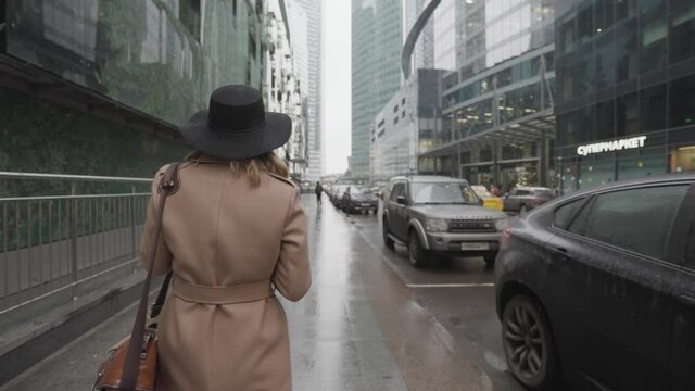 Elegant young lady wearing brown warm coat and black hat walking along high skyscrapers on a cold autumn day. Action. Female tourist holding camera outdoors in a modern city street. 