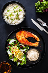 Fried salmon steak with basmati rice, mayonnaise and mix of vegetables served on black plate on wooden table
