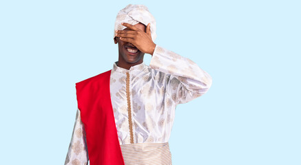 African handsome man wearing tradition sherwani saree clothes smiling and laughing with hand on face covering eyes for surprise. blind concept.