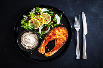 Fried salmon steak, mayonnaise and mix of vegetables served on black plate on wooden table
