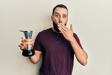 Young man with beard holding coffee maker with filter covering mouth with hand, shocked and afraid...