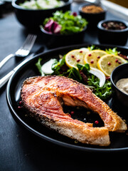 Fried salmon steak, mayonnaise and mix of vegetables served on black plate on wooden table
