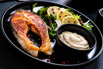 Fried salmon steak, mayonnaise and mix of vegetables served on black plate on wooden table

