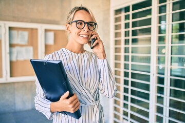 Young blonde businesswoman holding folder and talking on the smartphone at the city.