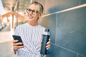 Young blonde businesswoman smiling happy using smartphone and drinking bottle of water at the city.