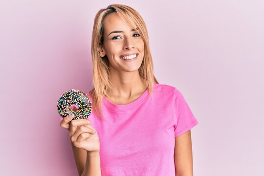 Beautiful blonde woman holding donut looking positive and happy standing and smiling with a confident smile showing teeth