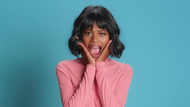 Impressed beautiful dark skinned woman looks with shocked surprised expression and says wow gestures as sees incredible poses for video against blue background. People emotions and lifestyle concept.