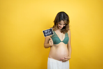 Young beautiful brunette woman pregnant expecting baby over isolated yellow background smiling and holding blackboard with new baby word message