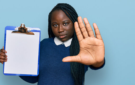 Young black woman with braids holding clipboard with blank space with open hand doing stop sign with serious and confident expression, defense gesture