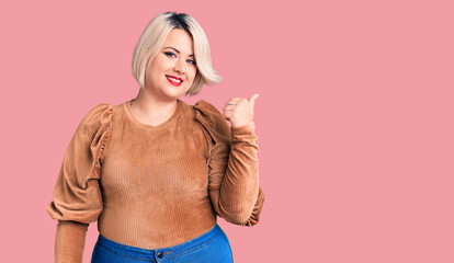 Young blonde plus size woman wearing casual sweater smiling with happy face looking and pointing to the side with thumb up.