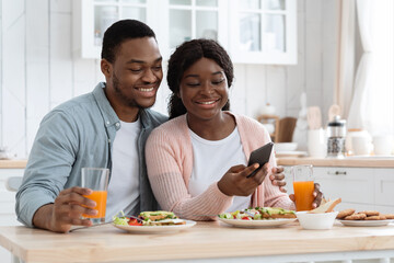 Obraz na płótnie Canvas Happy African American Spouses Using Smartphone While Having Breakfast In Kitchen