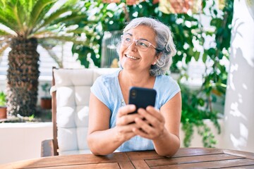 Middle age woman with grey hair smiling happy relaxing sitting at the terrace at home using...