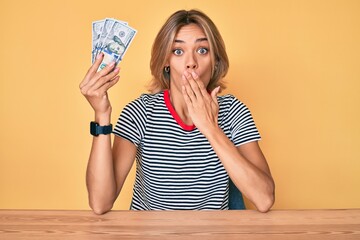 Beautiful caucasian woman holding 100 dollars banknotes covering mouth with hand, shocked and...