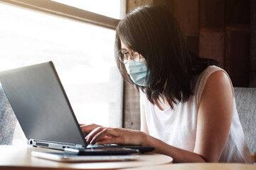 Asian young woman is wearing mask prevent for civid-19 and online working with laptop in office. She is searching information on internet at coffee shop. Businesswoman work with technology concept.