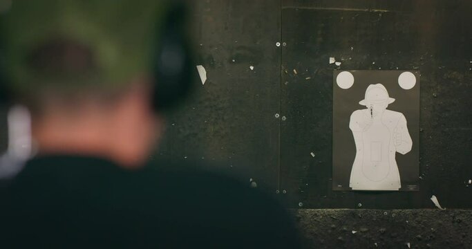 Rear view of a defocused man wearing ear protection aiming a pistol and shooting at a target at an indoor firing range.