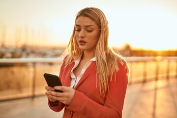 Young blonde businesswoman with serious expression using smartphone at the city.