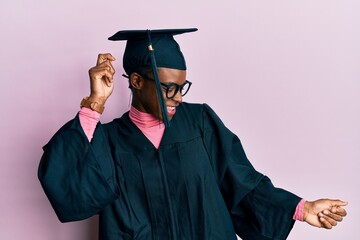 Young african american girl wearing graduation cap and ceremony robe dancing happy and cheerful,...
