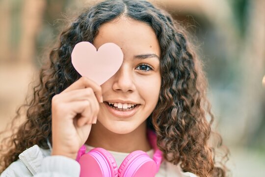 Hispanic teenager girl using headphones holding heart over eye at the city.