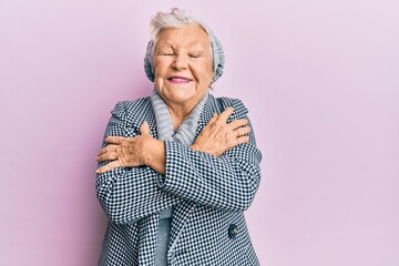 Senior grey-haired woman wearing wool winter sweater hugging oneself happy and positive, smiling...