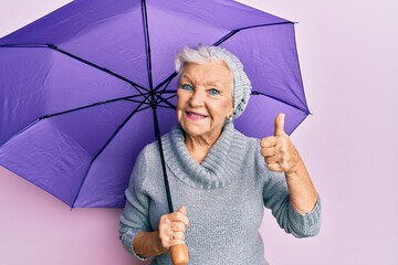 Senior grey-haired woman holding purple umbrella smiling happy and positive, thumb up doing...