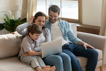 Smiling parents relax on sofa in living room with small 7s son child have fun browsing using modern...