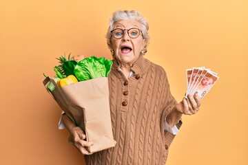 Senior grey-haired woman holding groceries and israel shekels banknotes celebrating crazy and...