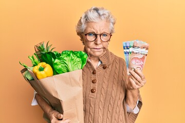 Senior grey-haired woman holding groceries and canadian dollars skeptic and nervous, frowning upset because of problem. negative person.