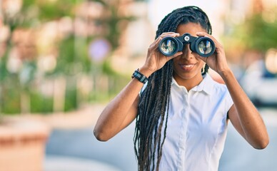 Young african american woman smiling happy looking for new opportunity using binoculars at the city.